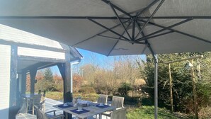 A covered outdoor dining area with a table and chairs, a large umbrella, and a view of the surrounding garden and trees. - Modern Holiday Home in Bazel With Garden (Kruibeke)
