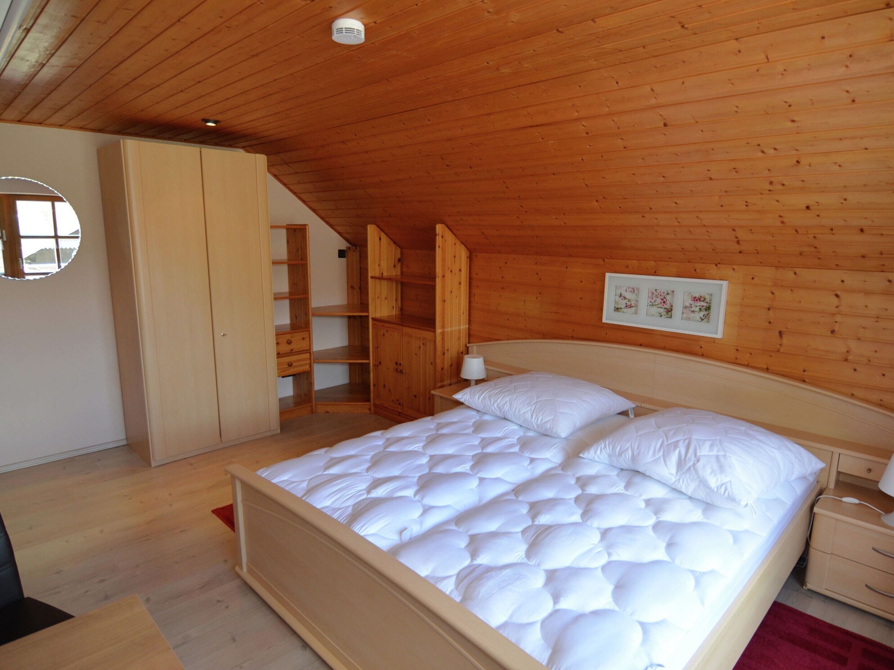 Ein Schlafzimmer mit Holzbalkendecke, einem Bett mit weißer Bettwäsche, einem Nachttisch, einer Kommode, einem Kleiderschrank und einem gerahmten Bild an der Wand.
