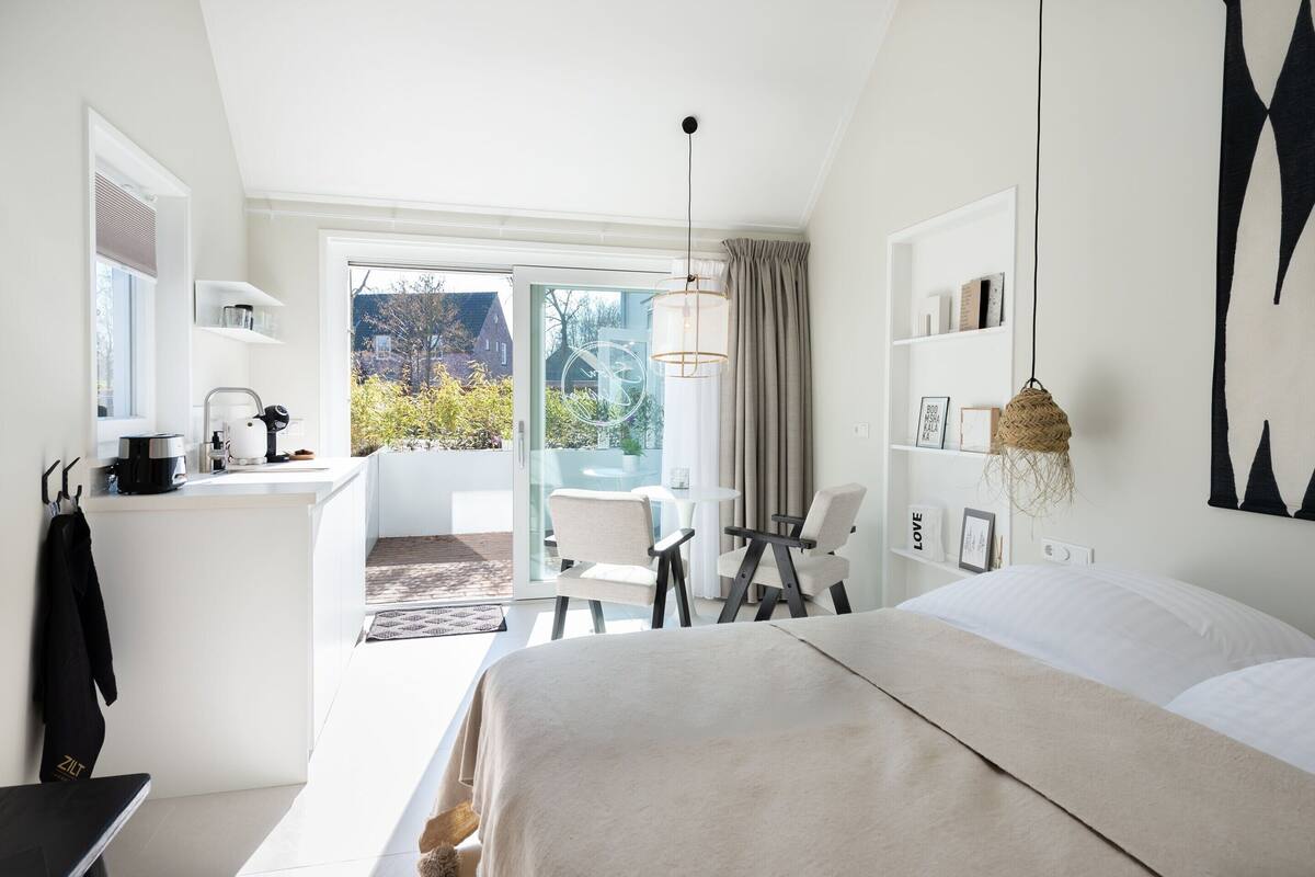 Una habitación de hotel moderna con una cama grande, un escritorio pequeño con cafetera, una silla y vistas al exterior a través de una puerta de cristal.