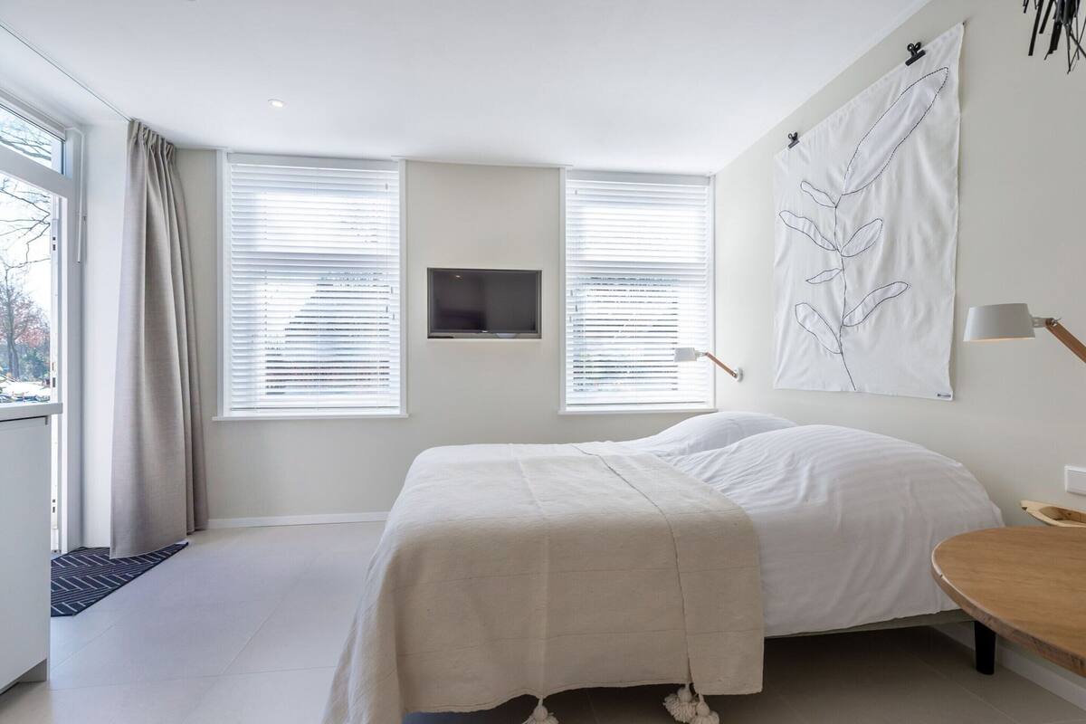 Un dormitorio moderno con una cama, una mesita de noche, un televisor montado en la pared y una ventana con persianas.