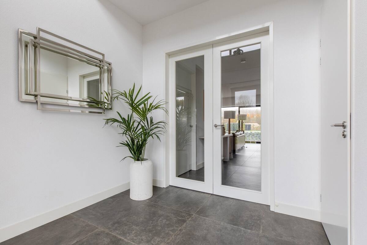 A modern hallway with a potted plant, a mirror, and a door leading to a room with a bed and a lamp.
