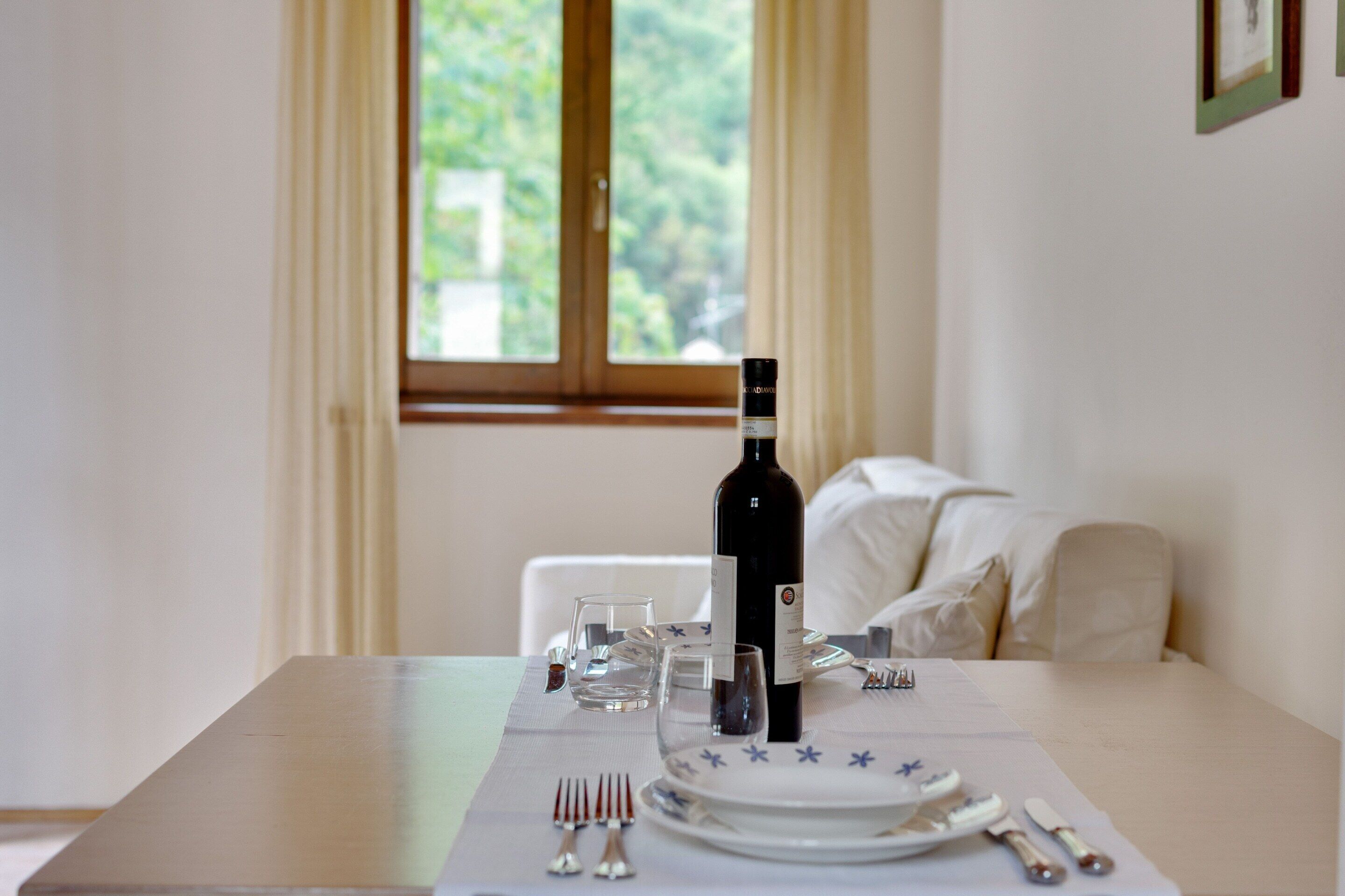 A dining table set with plates, glasses, cutlery, and a bottle of wine in a room with a window and curtains.