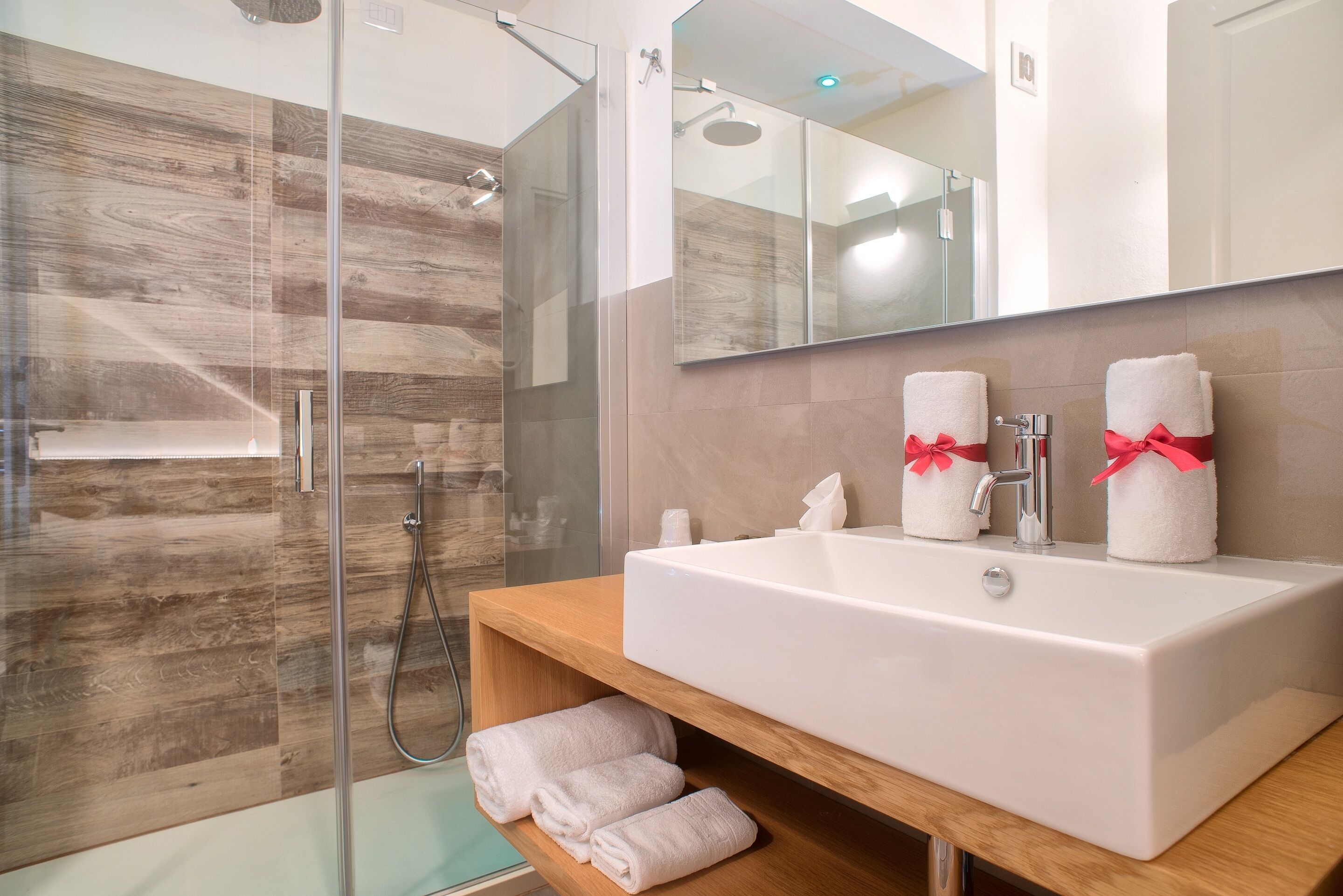 Une salle de bain avec une douche à parois de verre, un meuble-lavabo en bois avec un lavabo blanc et deux serviettes ornées de rubans rouges.