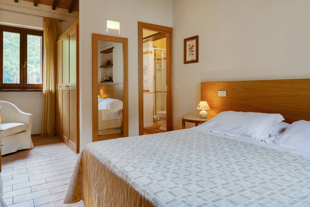 A bedroom with a bed, a chair, a wooden wardrobe, and a bathroom visible through an open door.