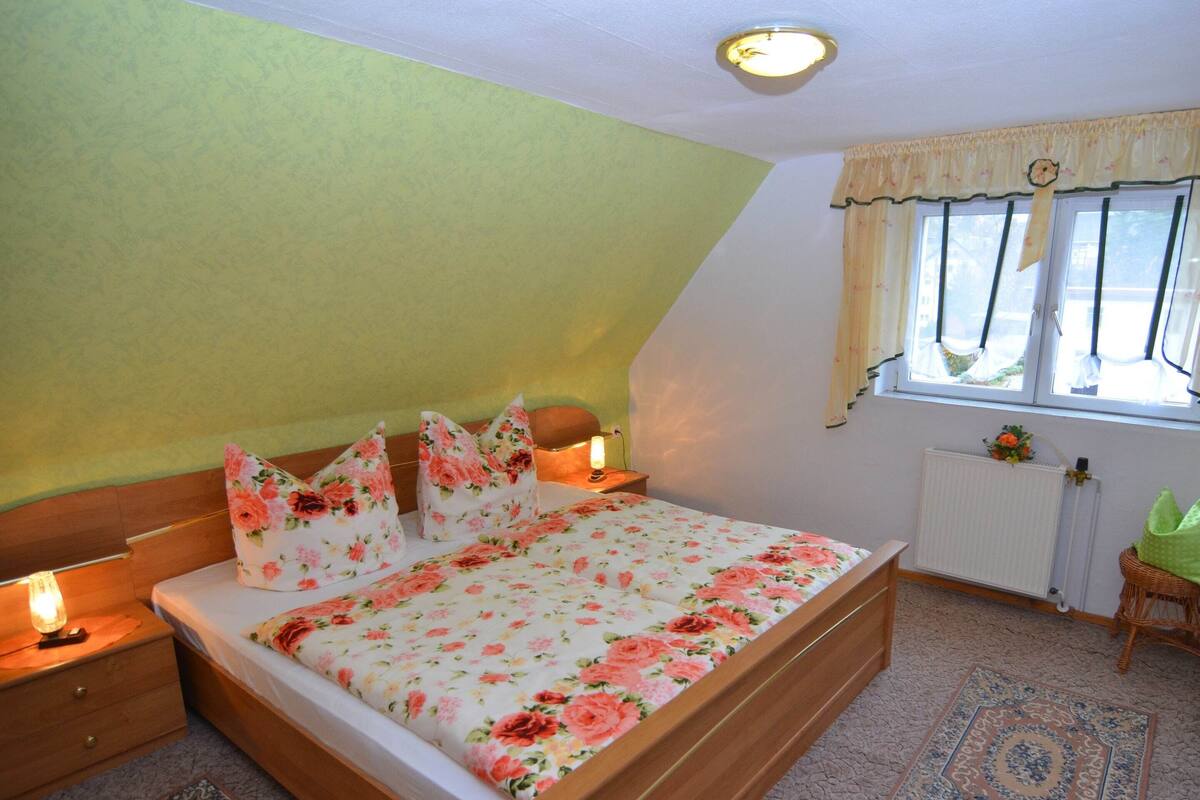 A bedroom with a wooden bed, floral bedding, a nightstand with a lamp, a window with curtains, and a radiator.