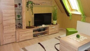 A cozy room with a wooden cabinet, a flat-screen TV, a small green table, and a window with green curtains. - Cozy Apartment in Lichtenhain Germany With Garden (Sebnitz)