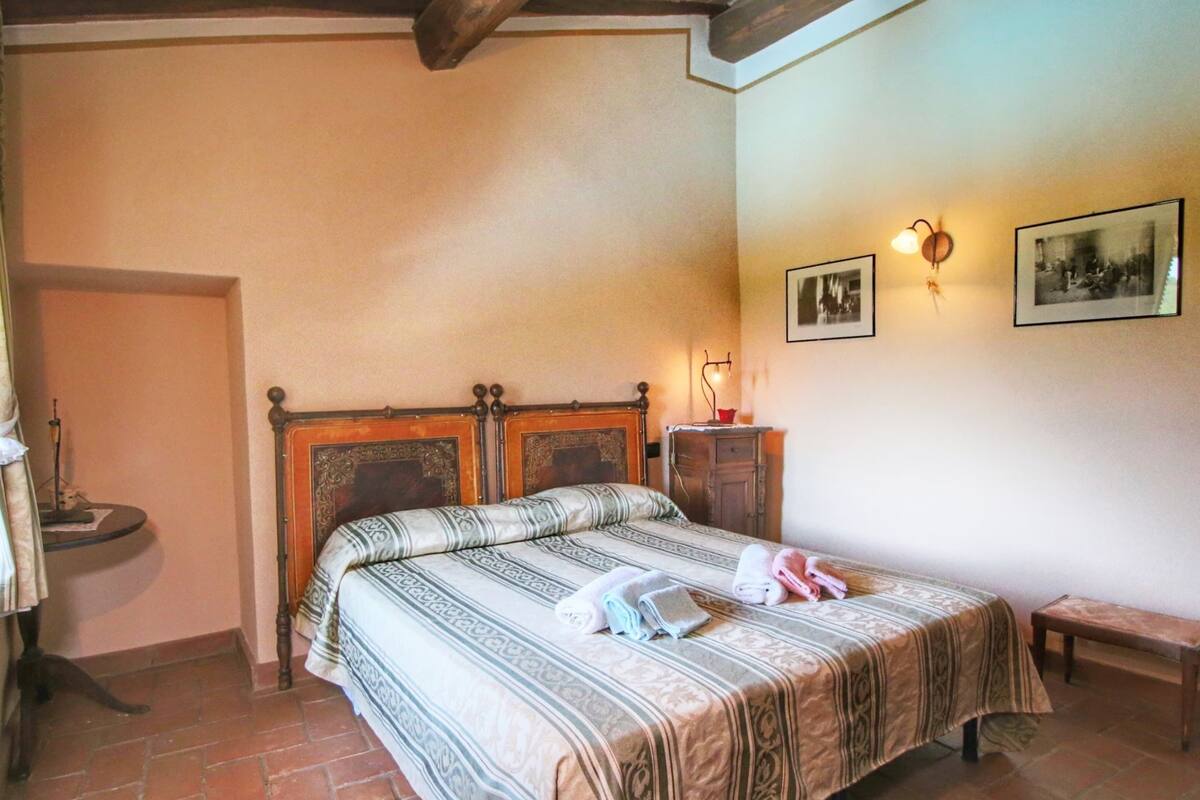 Un dormitorio con cama doble, cabecera de madera, mesita de noche con lámpara, un pequeño banco y dos cuadros enmarcados en la pared.