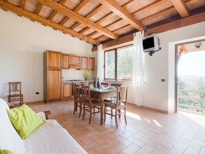 Une cuisine chaleureuse avec des meubles en bois, un coin repas avec des chaises, un téléviseur fixé au mur et une fenêtre donnant sur l’extérieur.