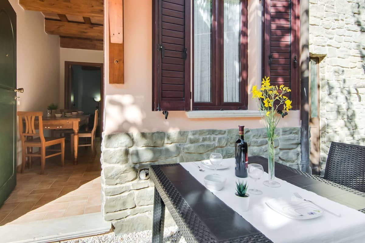 A stone-walled outdoor dining area with a table set for two, a bottle of wine, and a vase of yellow flowers.
