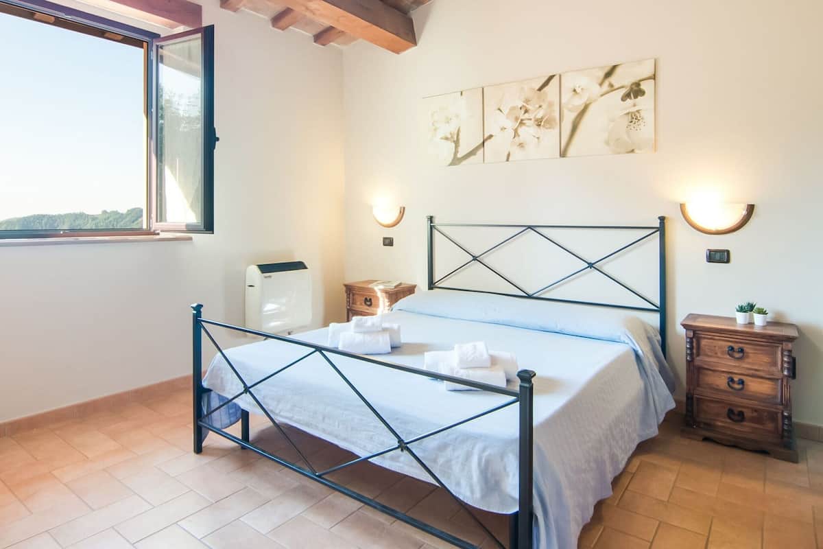 A bedroom with a bed, a nightstand, a wall-mounted heater, and a window with a view.