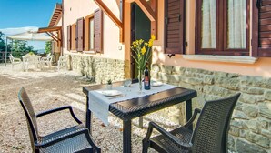 Une terrasse avec une table et des chaises, un mur en pierre, des poutres en bois et des fenêtres avec des volets.