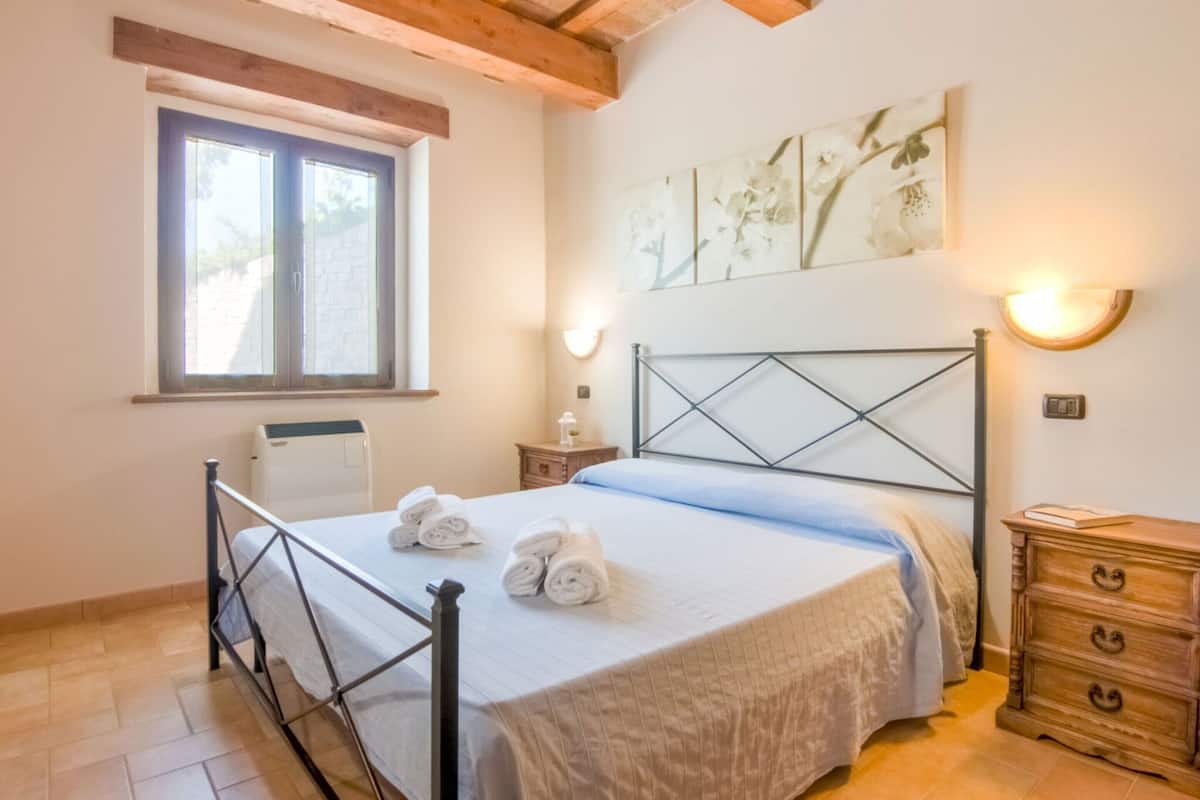 A bedroom with a bed, a window, a radiator, a wall-mounted light, and a wooden dresser.