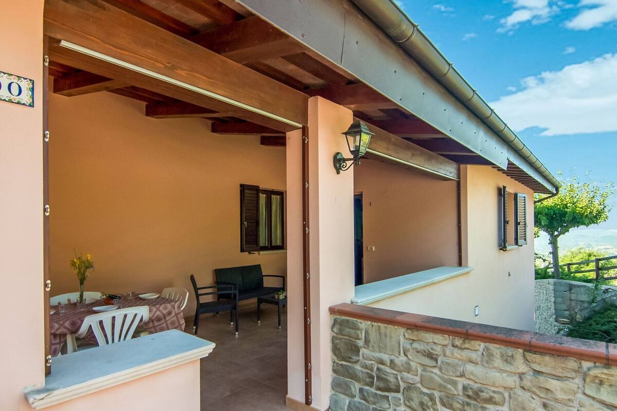 A covered outdoor seating area with a table and chairs, a stone wall, and a view of the landscape.