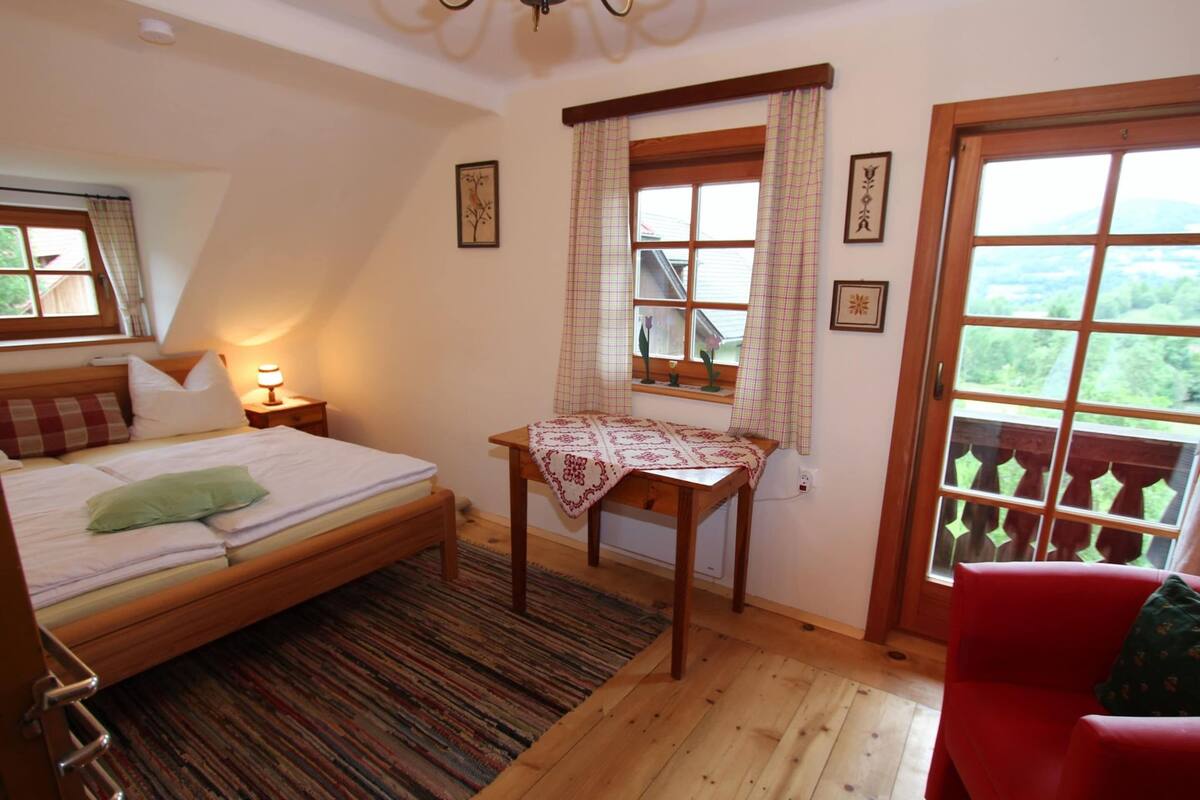 A bedroom with a bed, a small table with a tablecloth, a chair, and a window with curtains.