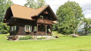 A wooden house with a sloped roof and a balcony, surrounded by a grassy yard and trees.