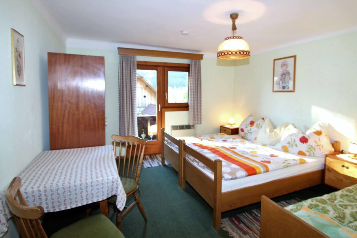 A bedroom with a bed, a wooden chair, a table with a patterned tablecloth, a nightstand with a lamp, and a window with curtains.