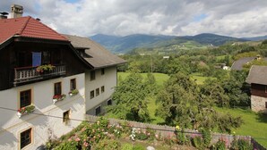 A traditional chalet with a balcony, surrounded by lush greenery and a mountainous backdrop.