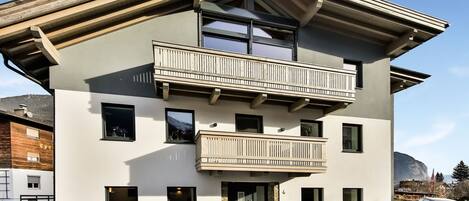 A modern two-story building with a balcony, a driveway, and a mountain backdrop.