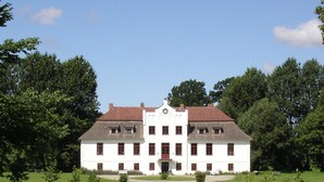 Ein großes, weißes Gebäude mit einem zentralen Eingang, umgeben von einer gepflegten Rasenfläche und Bäumen.