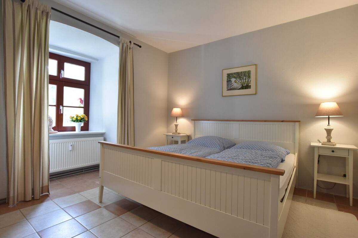 A bedroom with a bed, two bedside tables with lamps, a window with curtains, a radiator, and a framed picture on the wall.