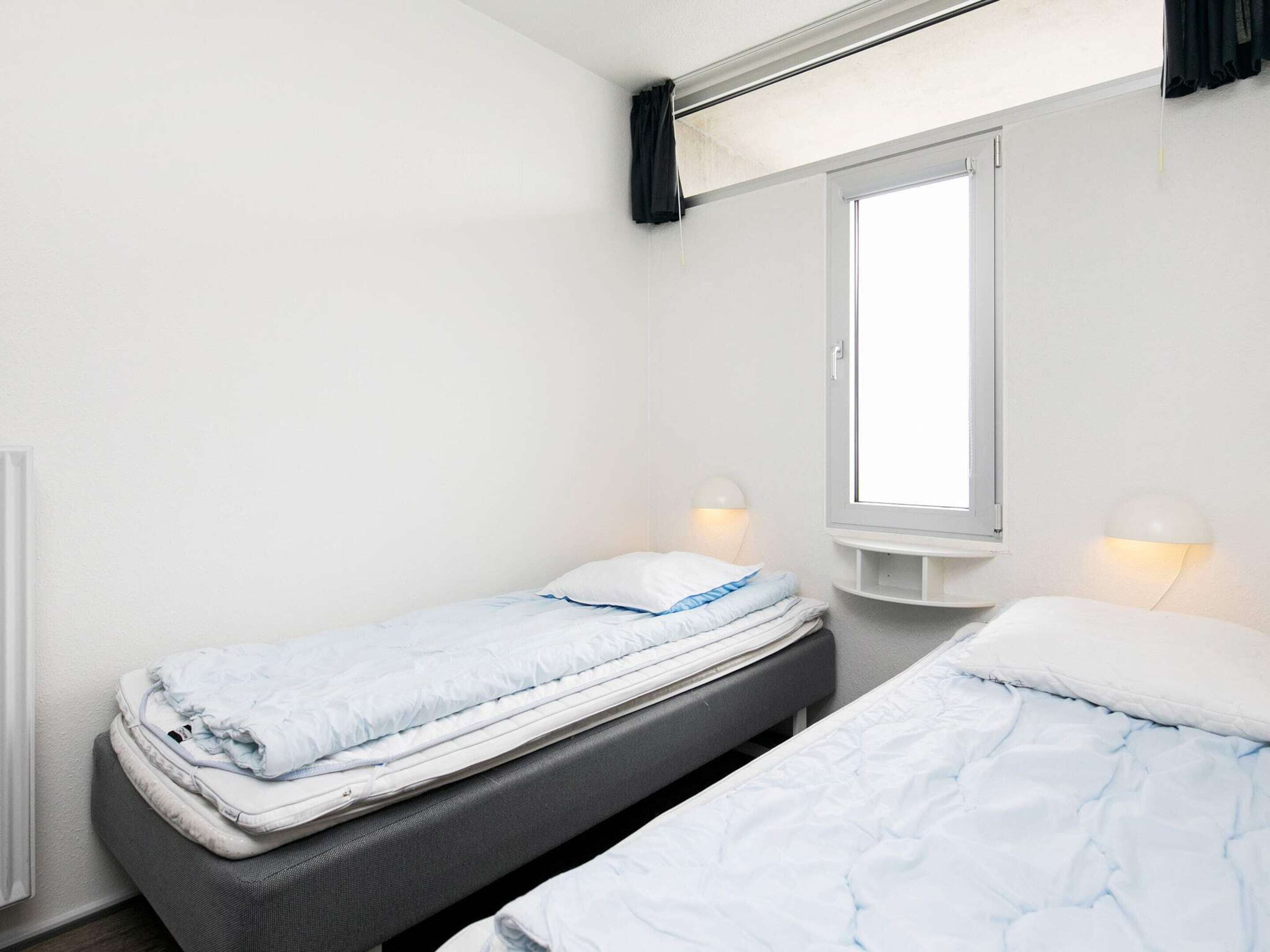 Une petite chambre propre, avec deux lits simples, une fenêtre avec des rideaux et des lampes de lecture fixées au mur.