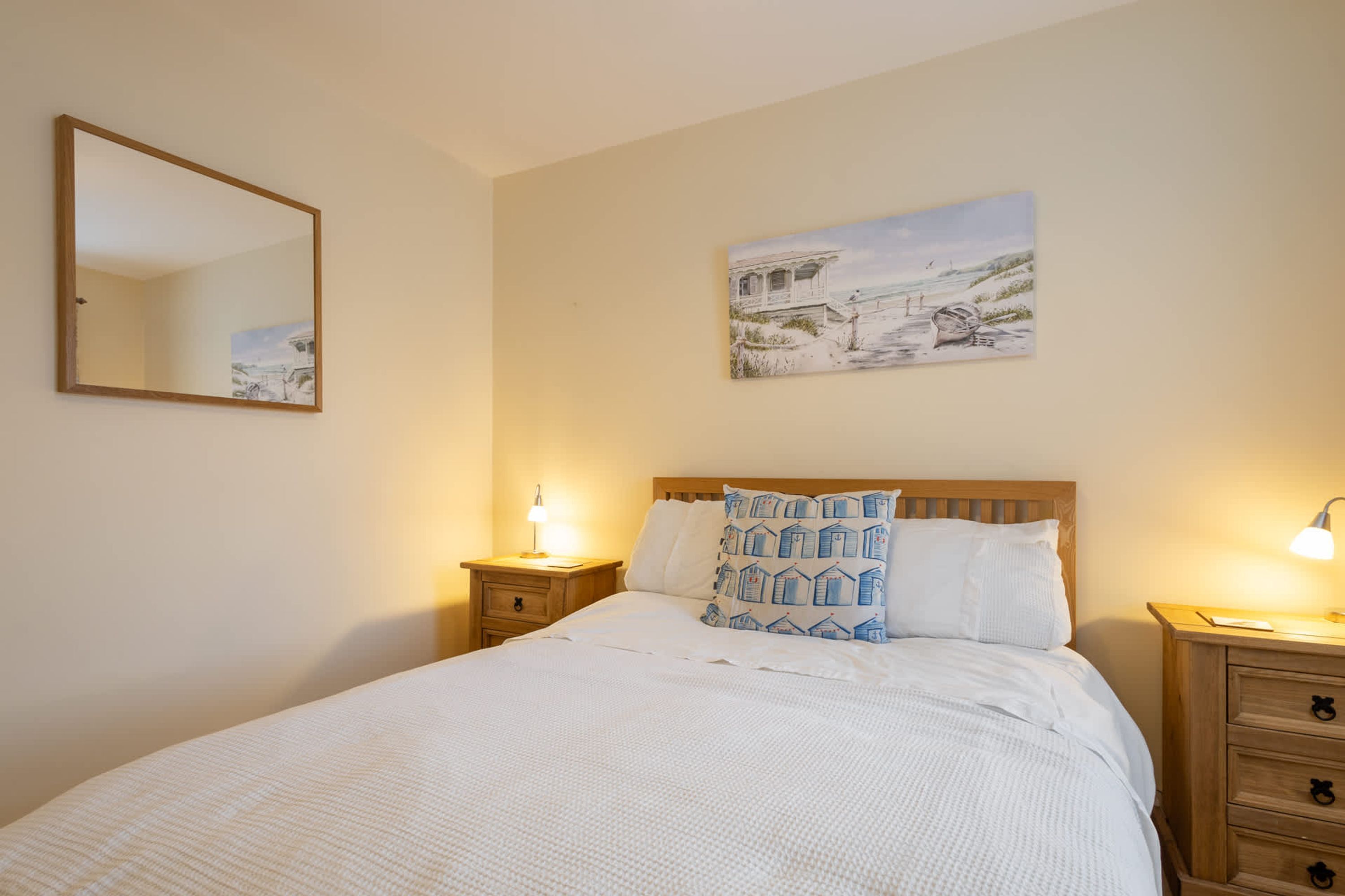 Un lit bien fait, avec une literie blanche et des oreillers à motifs, une table de chevet avec une lampe, et un tableau représentant un paysage côtier accroché au mur.