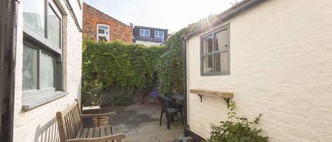 A small courtyard with a wooden bench, a table, and a chair. There is a white wall with a window and a door. A brick building is visible in the background.