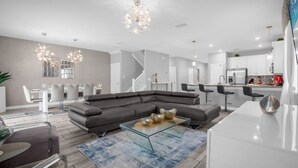 A modern living room with a sectional sofa, a glass coffee table, and a dining area with a white table and chairs. - Upstay Championsgate Resort Home w the Best Perks (Davenport)