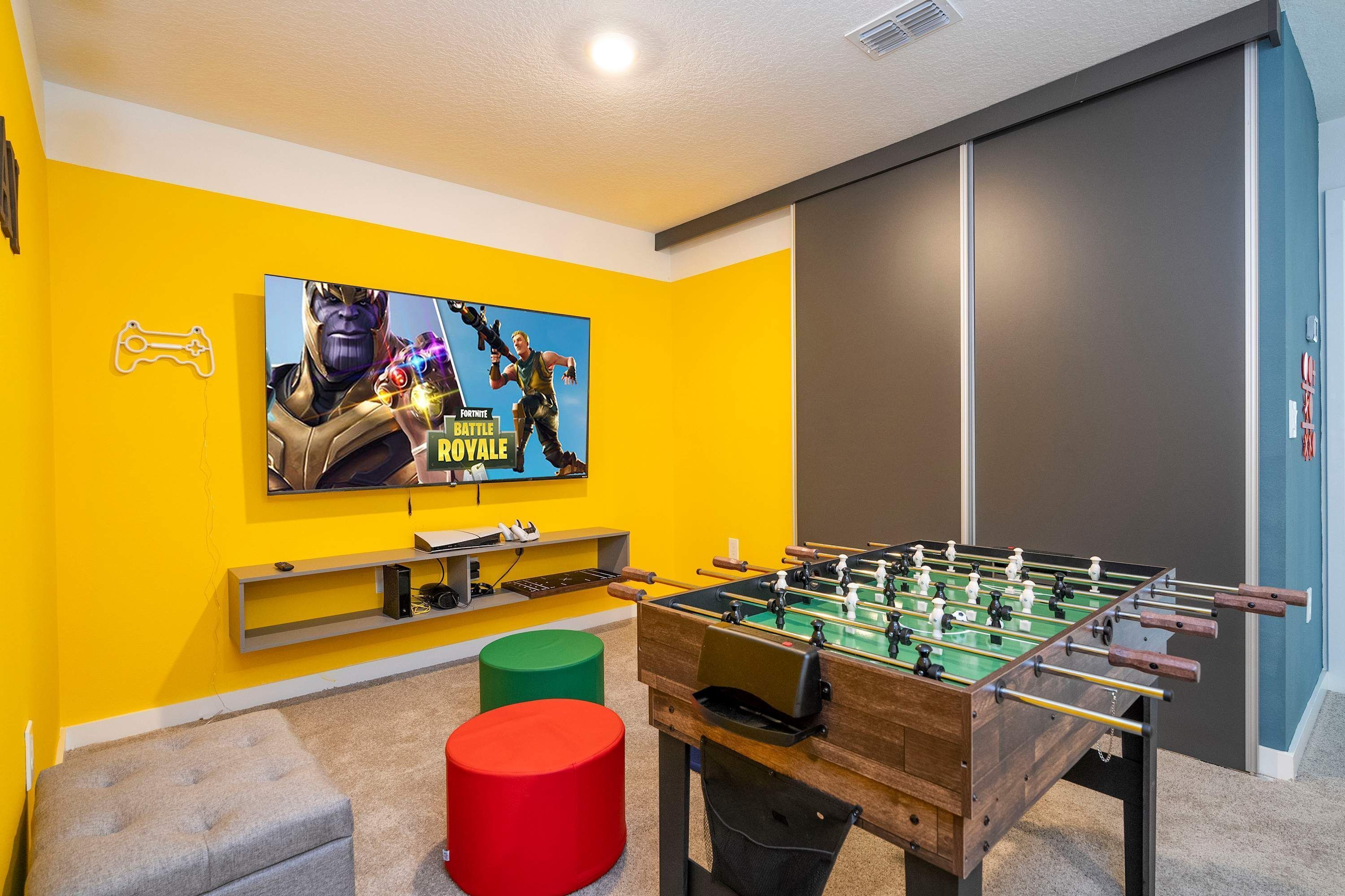 A room with a foosball table, a TV displaying a game poster, and a colorful wall.