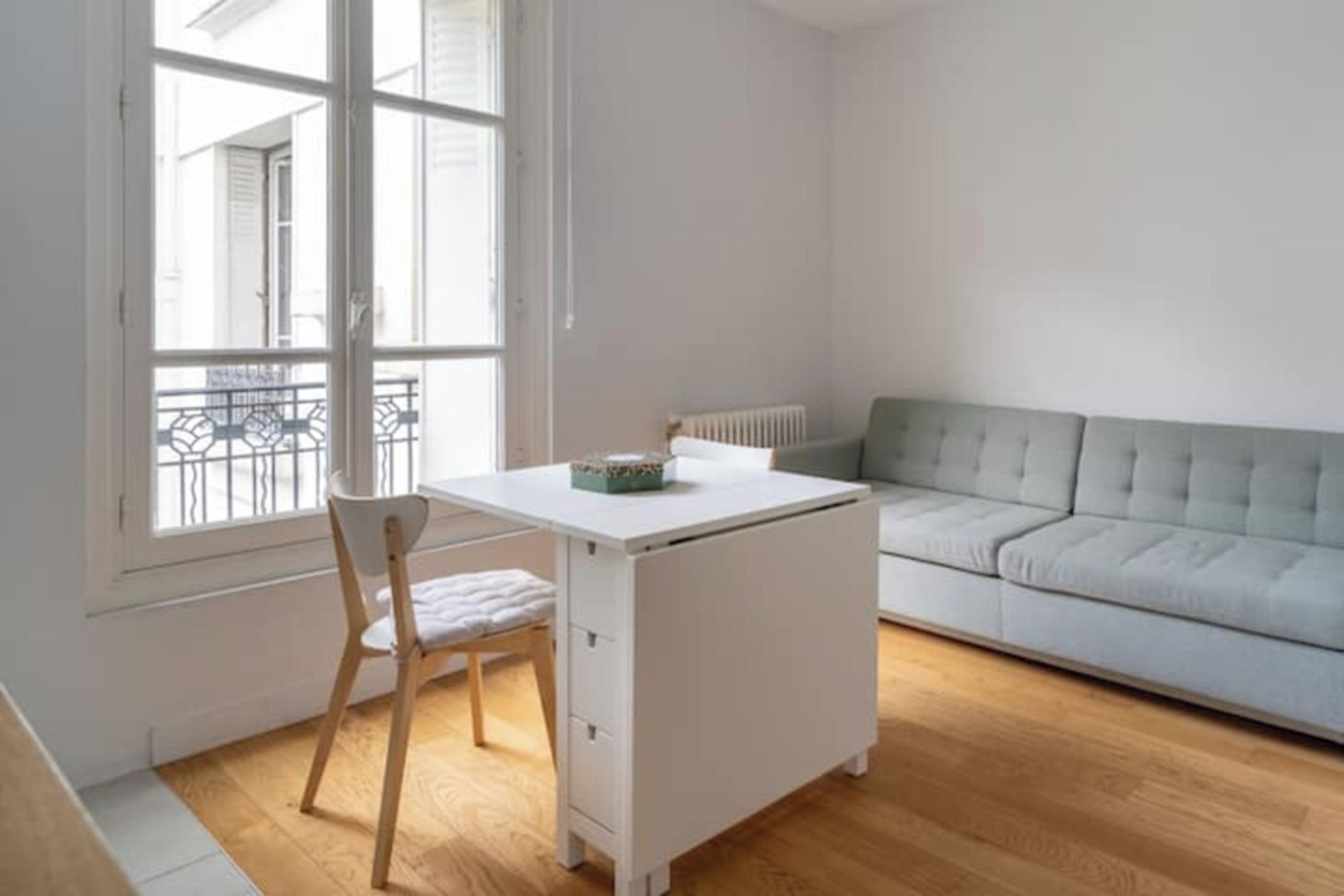 Una habitación con un escritorio blanco, un sofá gris y una ventana con vista a edificios.