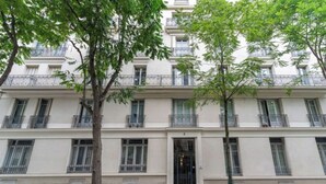 A multi-story building with balconies, a central entrance, and trees in front.
