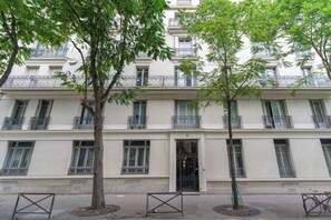 A multi-story building with balconies, a central entrance, and trees in front.