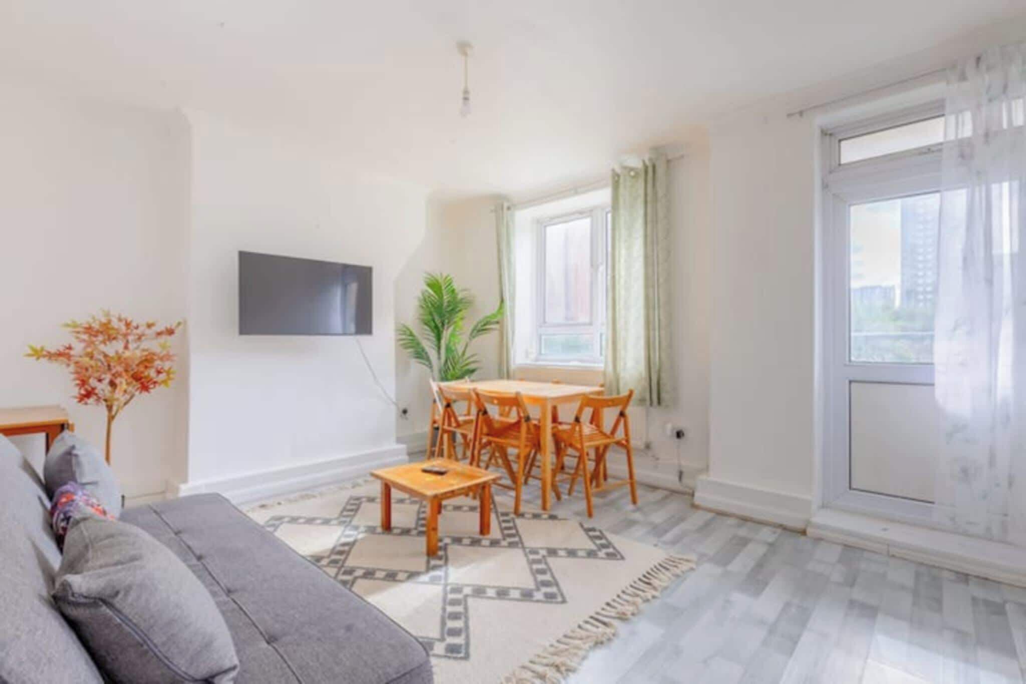 A living room with a sofa, a dining table with chairs, a TV mounted on the wall, and a large window with curtains.
