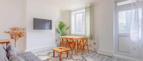 A living room with a sofa, a dining table with chairs, a TV mounted on the wall, and a large window with curtains.