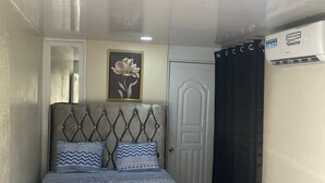 Un dormitorio con cabecera capitonada, cama con ropa de cama estampada, una obra de arte floral enmarco y una puerta blanca, además de un equipo de aire acondicionado.