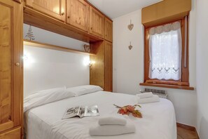 A neatly made bed with white linens, a magazine, and a towel on the bed. There is a window with a lace curtain and a radiator below it. Wooden cabinets are above the bed. - Wolf & Deer Apartment Ski-in Ski-out 250m (Sestriere)