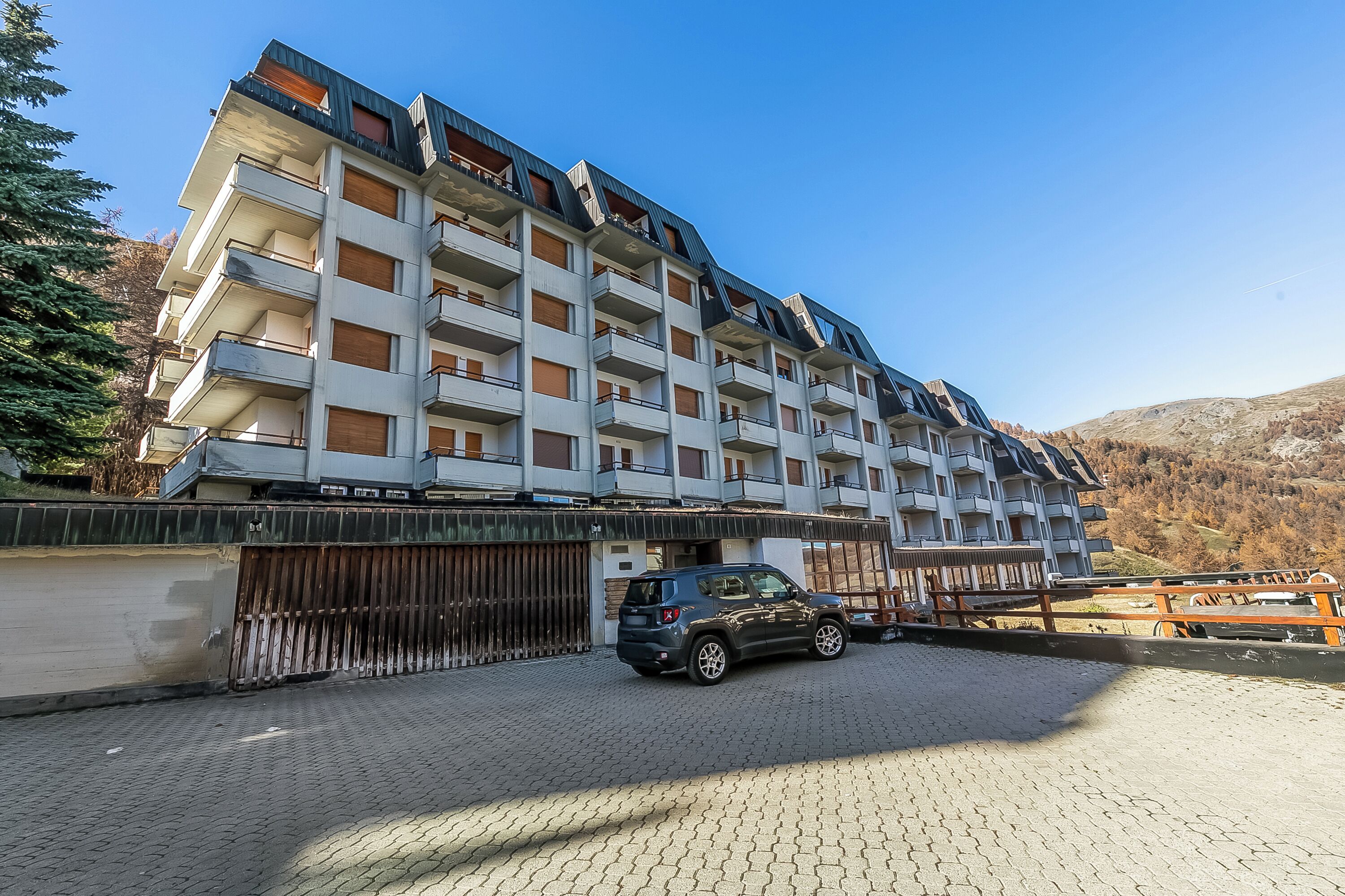 A multi-story hotel with balconies, a parking area, and a mountainous backdrop.
