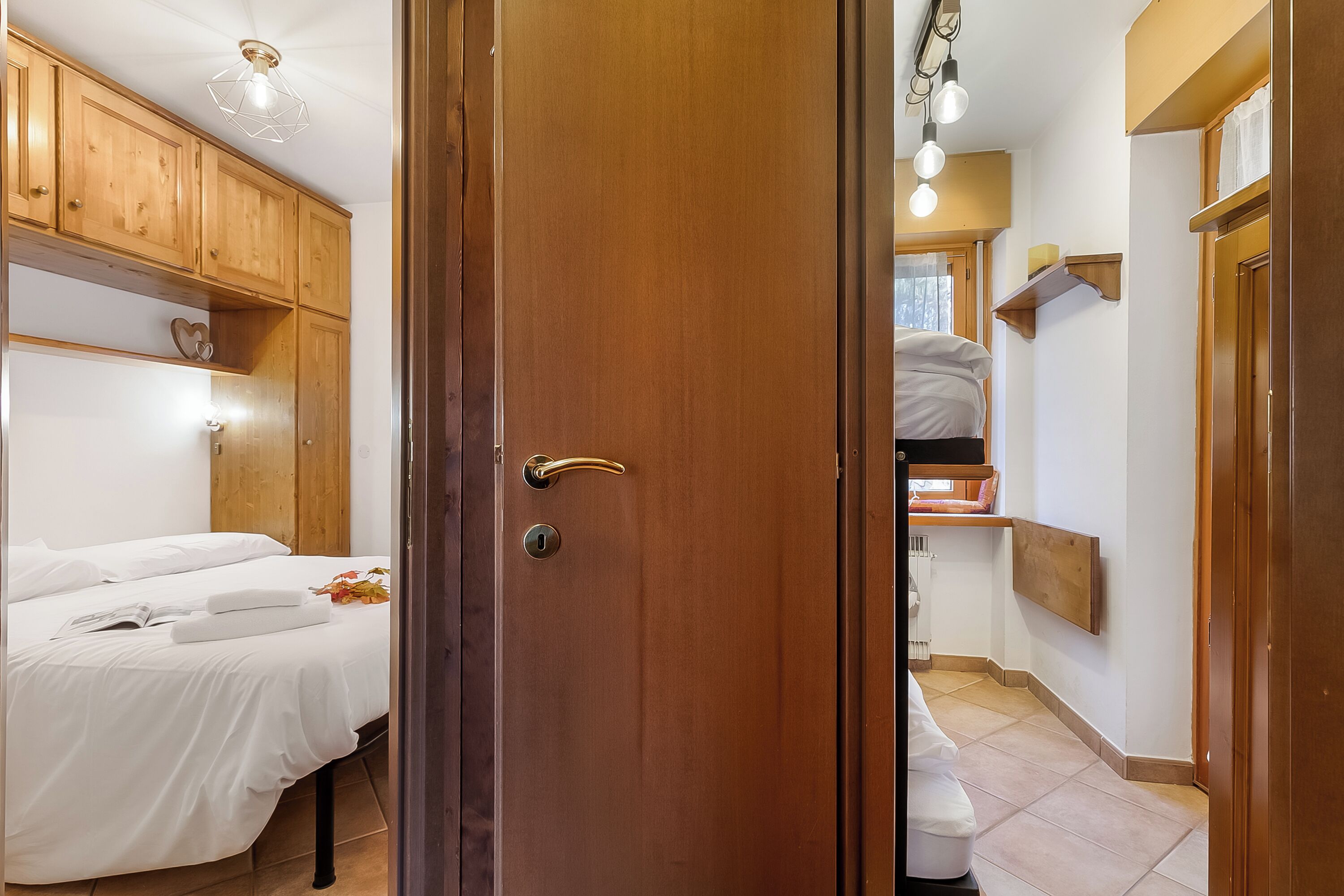 Hotellihuone, jossa on puinen vaatekaappi, valkoisilla lakanoiden peitetty sänky, yöpöytä, jossa on kirja ja kukka, sekä puinen hylly, jossa on sydämenmuotoinen koriste.