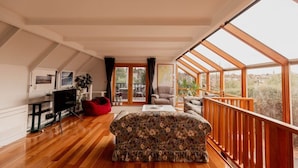 A spacious living room with a wooden floor, a floral sofa, a red bean bag, a TV, and a balcony with large windows. - Family Home in central Sandy Bay (Sandy Bay)