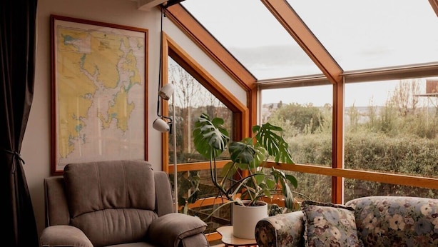 A room with a large window, a map on the wall, a brown chair, a floral sofa, and a potted plant. - Family Home in central Sandy Bay (Sandy Bay)