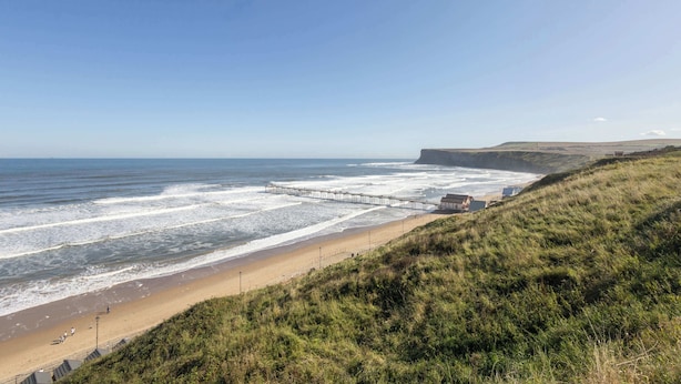 A coastal beach with a pier extending into the sea, grassy hills, and a clear sky. - Host & Stay | Garnet Street (Saltburn-by-the-Sea)