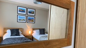 A hotel room with two beds, a wooden headboard, a nightstand with a lamp, and a mirror reflecting the room. There are framed pictures on the wall. - Luxury 2-bedroom apartment  in sleepy Filey on the North Yorkshire coast  (Filey)
