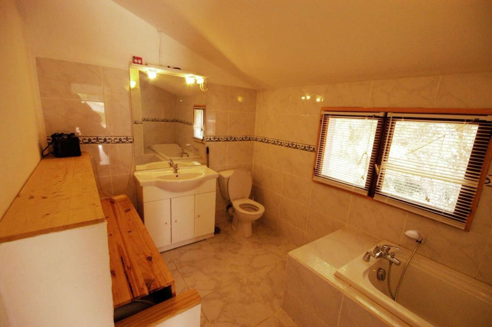 Une salle de bain comprenant des toilettes, un lavabo et une baignoire. Il y a une fenêtre avec des stores, un miroir au-dessus du lavabo et un banc en bois.