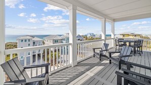 A covered outdoor deck with Adirondack chairs and a table, overlooking a beach and residential buildings. - Gulf Views w/ Pool, Firepit and Games (Port St. Joe)