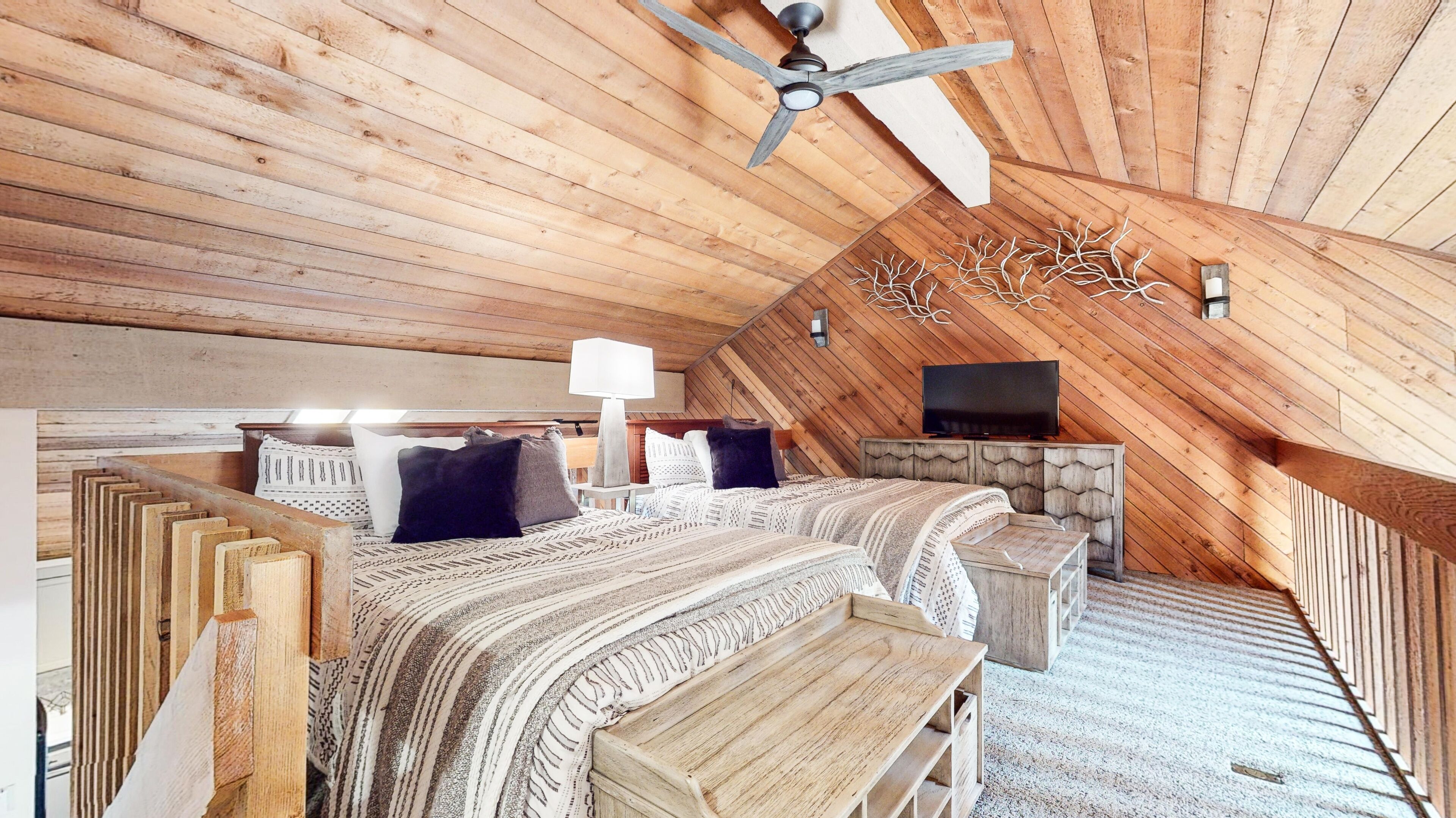 Une chambre aménagée dans les combles, avec des lambris en bois au plafond, un ventilateur de plafond, une télévision, une cheminée et un lit recouvert d’une literie à motifs.