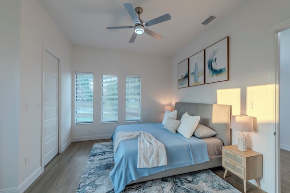 A bedroom with a bed, bedside table, ceiling fan, and artwork on the wall.