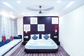 A modern hotel room with a large bed, a seating area with a sofa and pillows, a wall with framed artwork, and a ceiling light fixture. - Hide-in Udaipur (Udaipur)