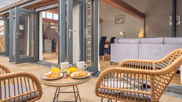 An open patio with wicker chairs, a breakfast table with croissants and juice, and a view into the interior of a modern home.