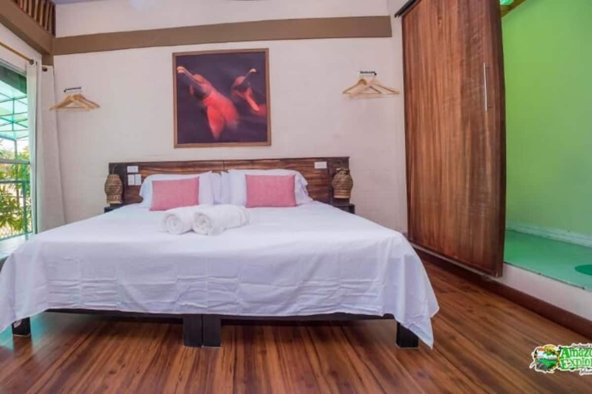 A neatly made bed with white linens and pink pillows, a wooden floor, a large window with curtains, and a painting on the wall.