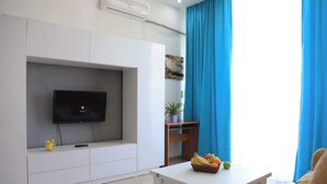 A modern living room with a flat-screen TV mounted on a white cabinet, a small table with a fruit bowl, and a blue curtain. - Kristal Absheron 110 (Baku)
